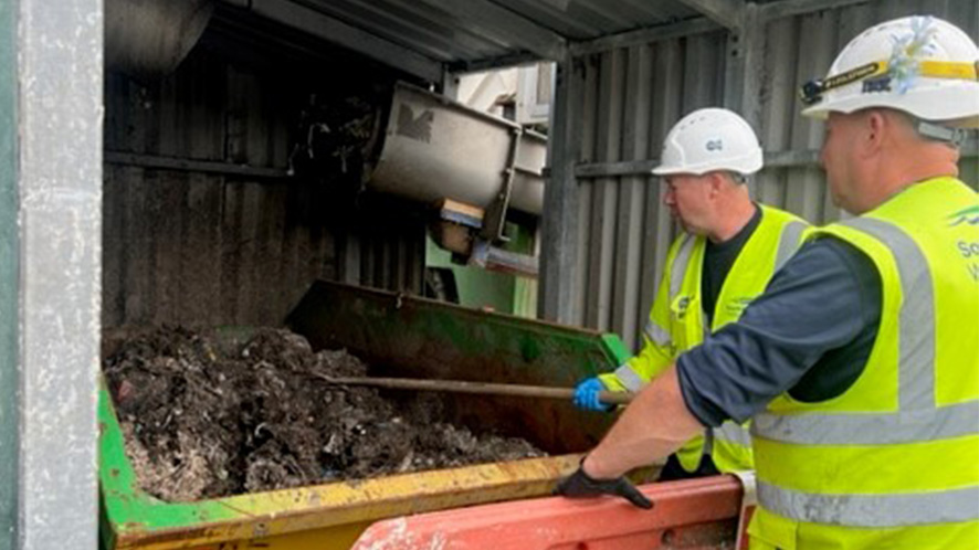 Two wastewater treatment works operatives rake the top of the skip where wipes collect to level out the discarded material